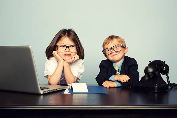 Two young kids in glasses and business clothes sitting at a desk with a laptop, folder and old rotary phone — playing at running their own business