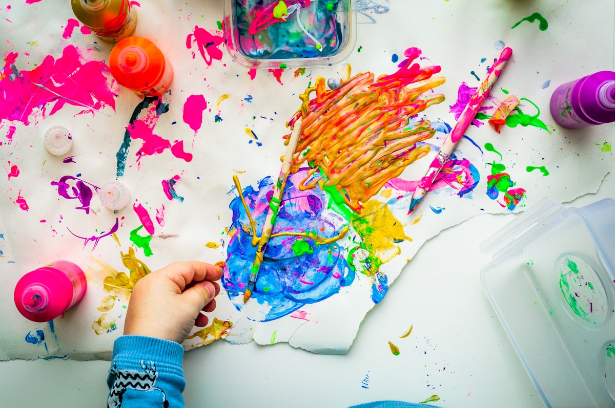 Child's hands deep in a gloriously messy finger-painting session — paint bottles and torn paper everywhere