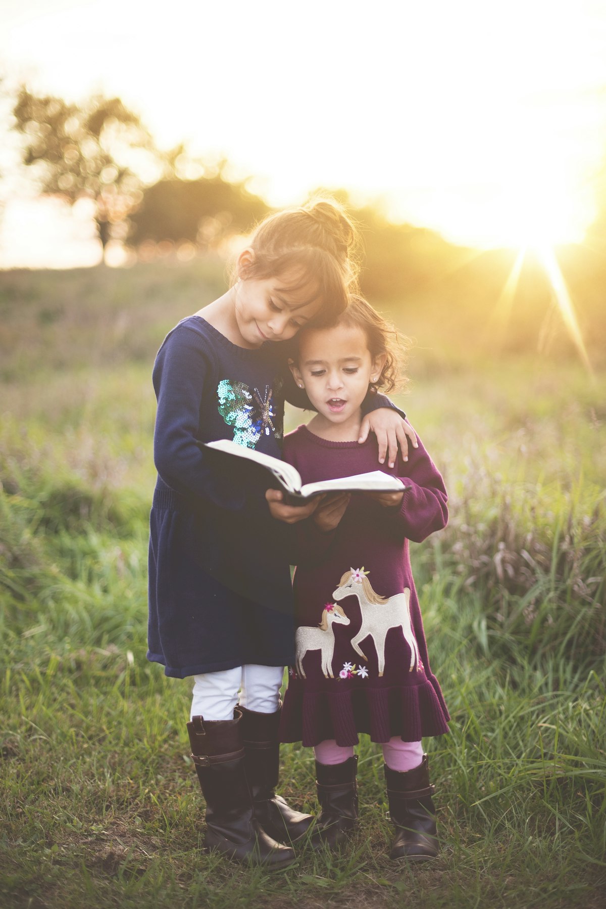 Two young sisters reading a book together in a golden field at sunset — self-chosen, unstructured time