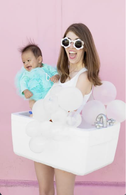 Woman holding a baby in a mint outfit, wearing a white tub costume covered in soap bubbles against a pink wall