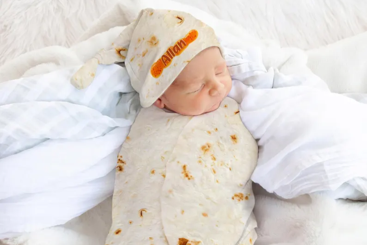 Newborn asleep in a tortilla-print swaddle and matching knotted hat, on a white fur blanket