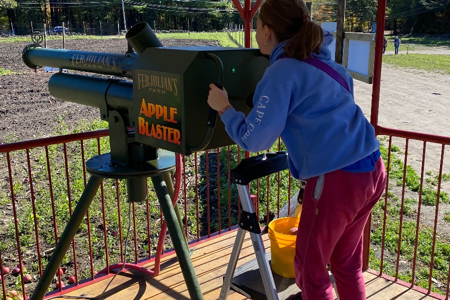A child leaning into the green Ferjulian’s Farm Apple Blaster on a covered deck, with a yellow bucket of apples beside her