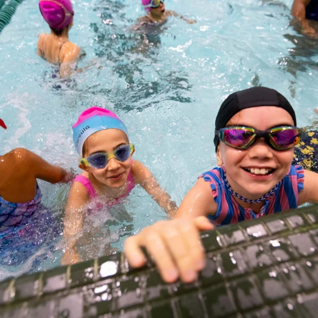 Children in swim caps and goggles smiling at the edge of an indoor pool at Asphalt Green