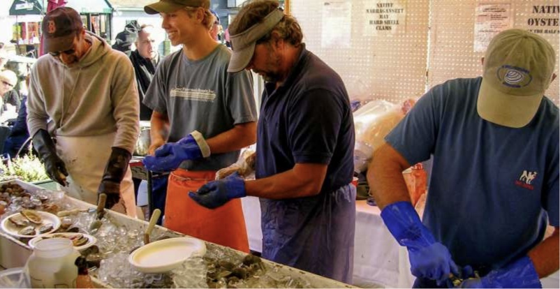 Shuckers opening oysters at a seafood stand, with blue gloves and aprons, at the Bowen’s Wharf Seafood Festival in Newport