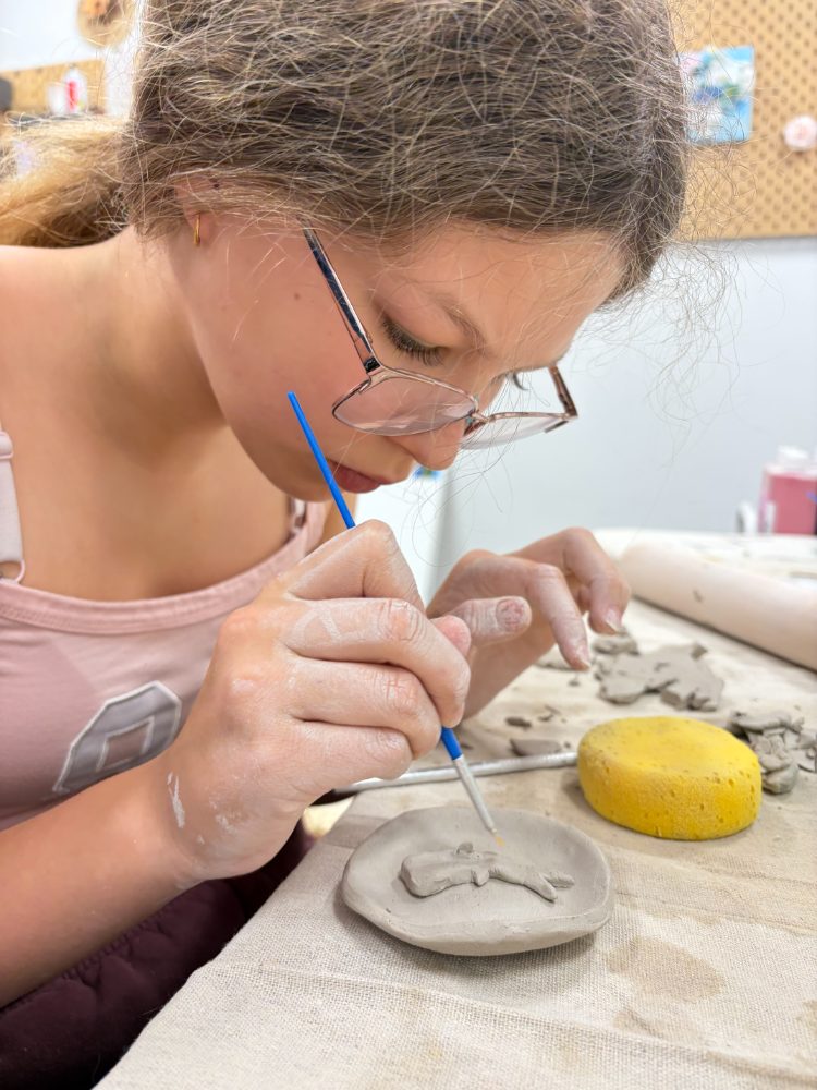 A girl painting a small clay figure onto a hand-built clay dish