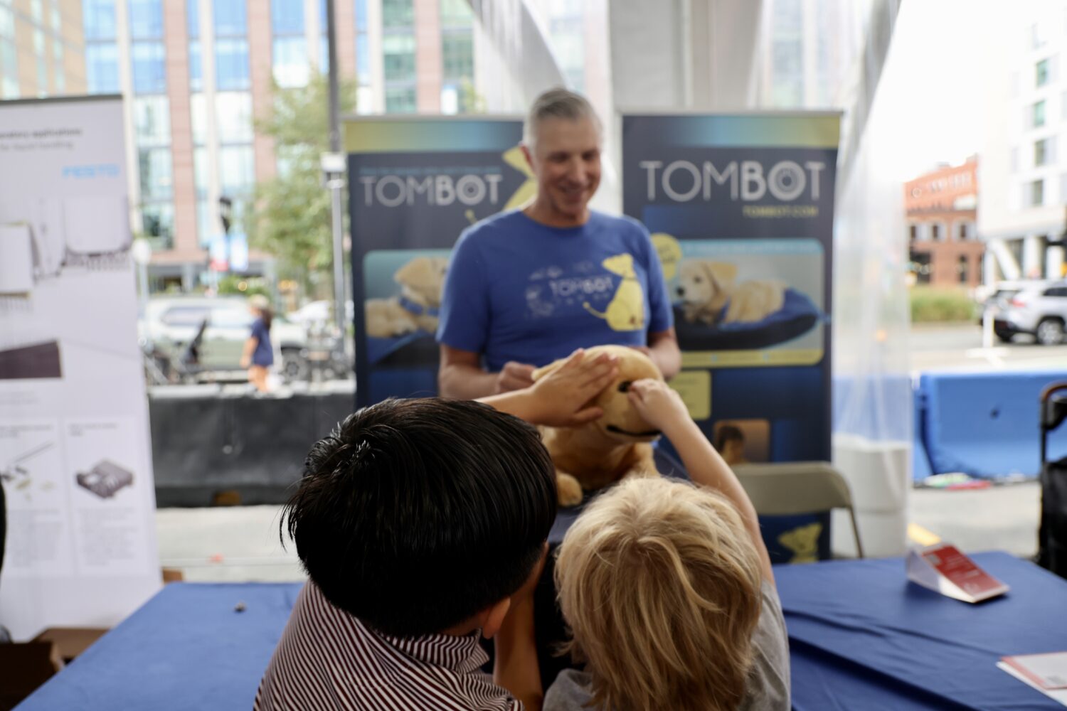 Two children reaching up to stroke a robotic golden retriever at a Tombot stand during RoboBoston in the Boston Seaport