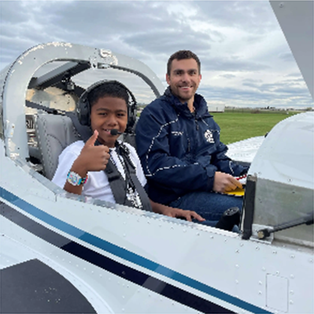 A smiling young boy in a headset gives a thumbs-up from the cockpit of a small airplane beside a volunteer pilot, on a Young Eagles flight